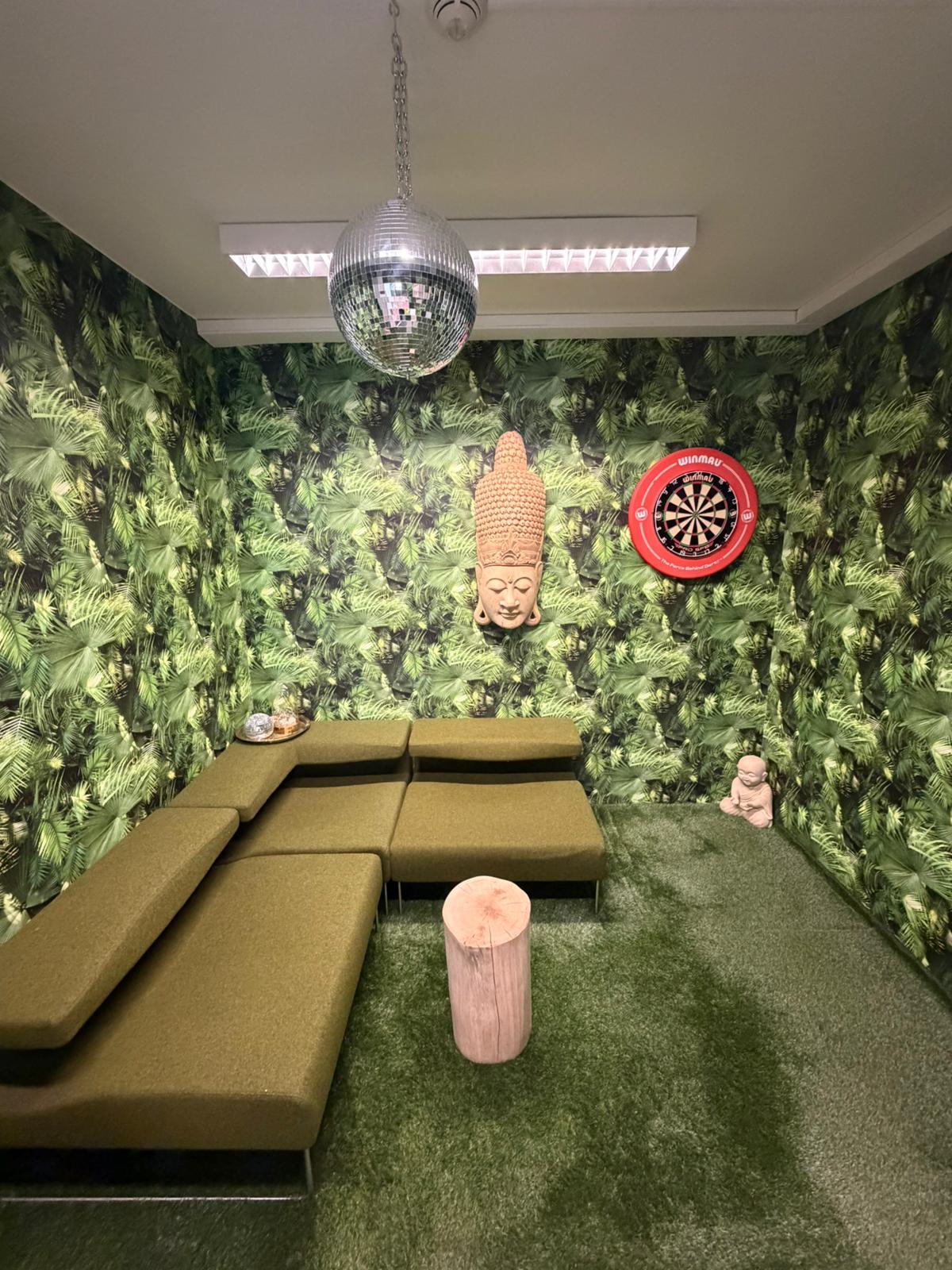 Eine kreative Jungle-Lounge mit Pflanzen-Tapete, olivgrünem Sofa, Holzstamm-Hocker, einer Discokugel und einer Dartscheibe
