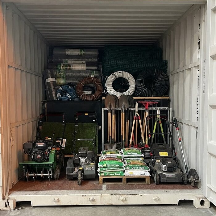 Lagercontainer eines Garten- und Landschaftsbaubetriebs mit Rasenmähern, Schaufeln, Rechen und Säcken auf einer Palette
