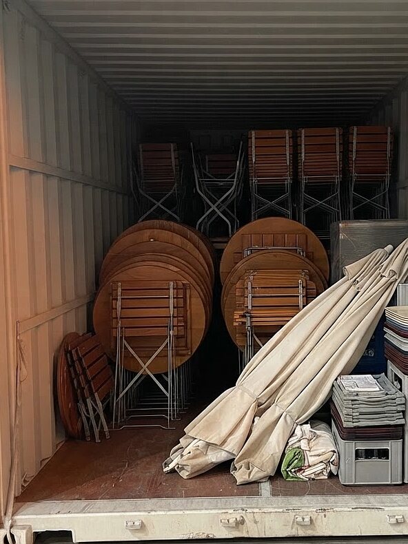 Lagercontainer eines Gastronomiebetriebs mit gestapelten Biertischen, Klappstühlen und zusammengelegten Sonnenschirmen