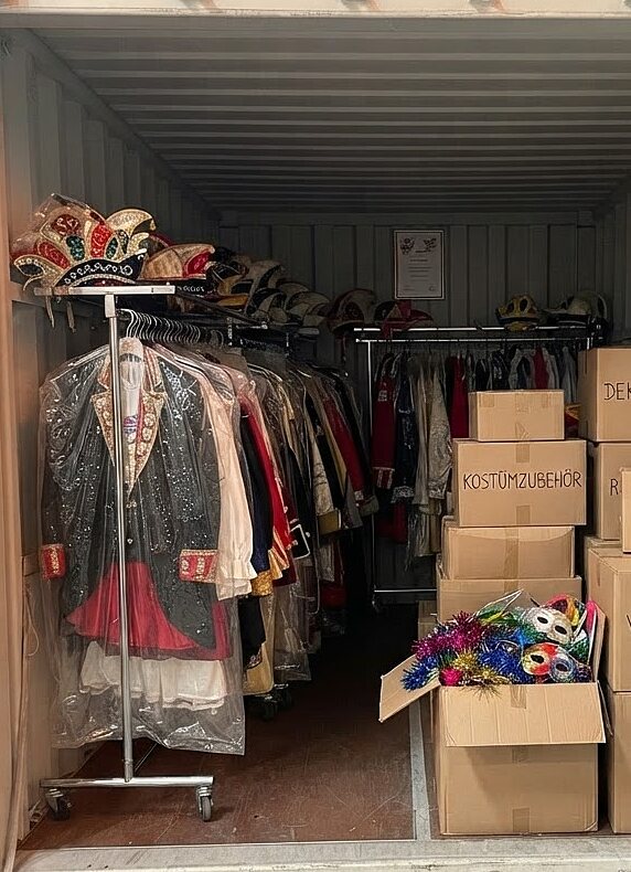 Lagercontainer eines Karnevalsvereins mit Kostümen auf einer Kleiderstange, Kopfbedeckungen und beschrifteten Kartons für Vereinsmaterial