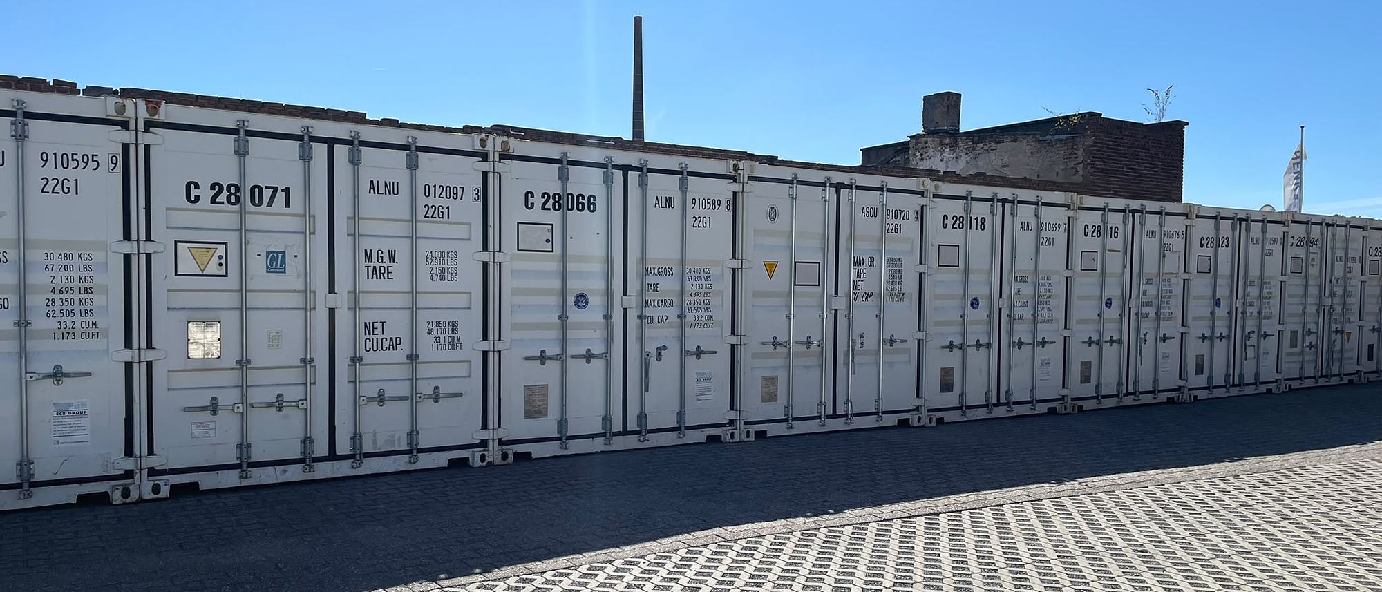 Der neue Selfstorage-Lagerpark von Henk in Düsseldorf: eine lange Reihe weißer Lagercontainer an einer breiten, gepflasterten Zufahrt unter blauem Himmel