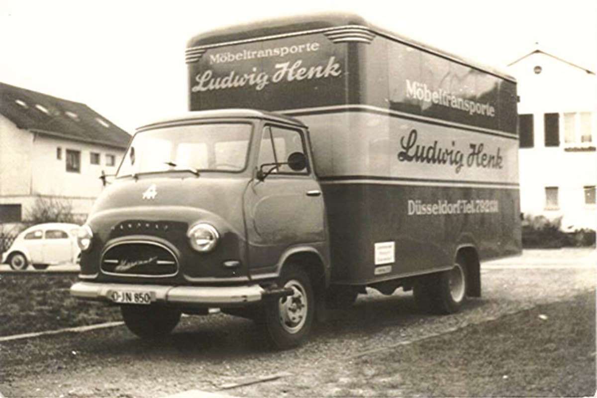 Ein altes Schwarz-Weiß-Foto eines kastenförmigen Transportwagens mit der Aufschrift Möbeltransporte Ludwig Henk, der an Häusern geparkt ist. Das Fahrerhaus ist rund mit einem Borgward-Logo, und der Lkw wirbt auf seiner Seite für Möbeltransporte in Düsseldorf.