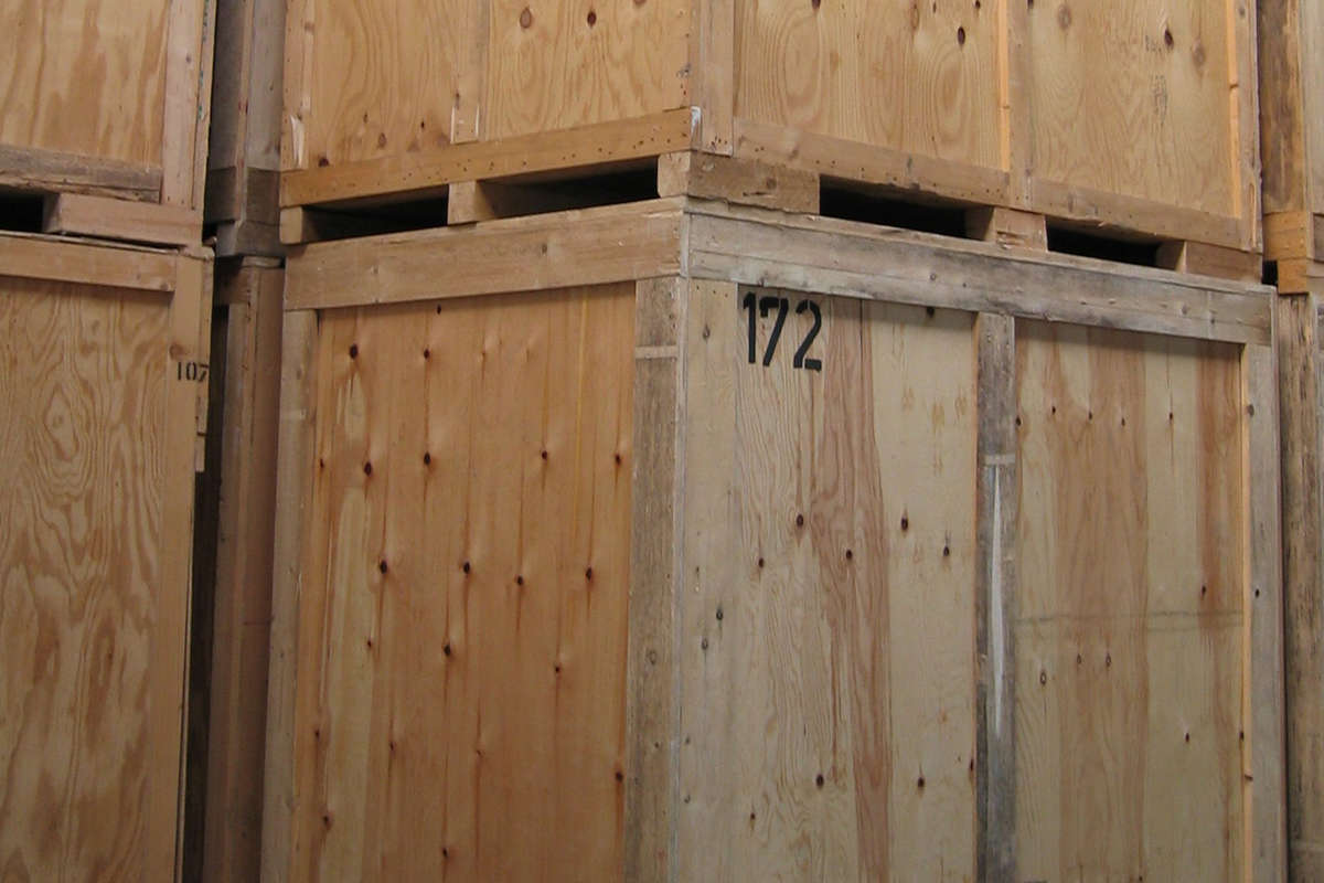 Mehrere große, gestapelte Holzkisten mit sichtbaren Nägeln und Maserungen; die vordere Kiste ist mit 172 in schwarzen Zahlen beschriftet. Die Kisten bestehen aus unbearbeitetem Sperrholz, und die Szene deutet auf ein Lagerhaus oder eine Lagereinrichtung hin.