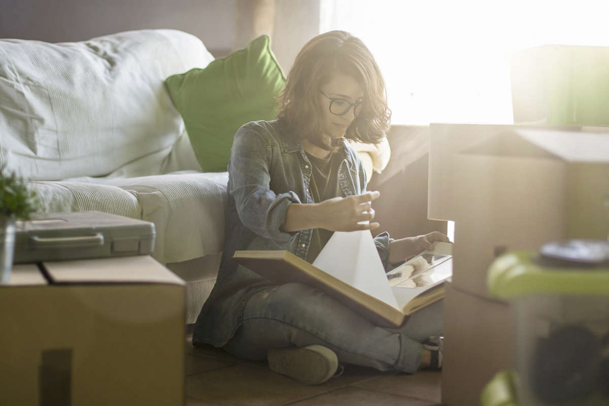 A woman wearing glasses and with shoulder-length hair is sitting cross-legged on the floor next to a couch, flipping through a photo album. In a sun-drenched room, she is surrounded by moving boxes, giving the impression that she is in the process of packing or unpacking her belongings.